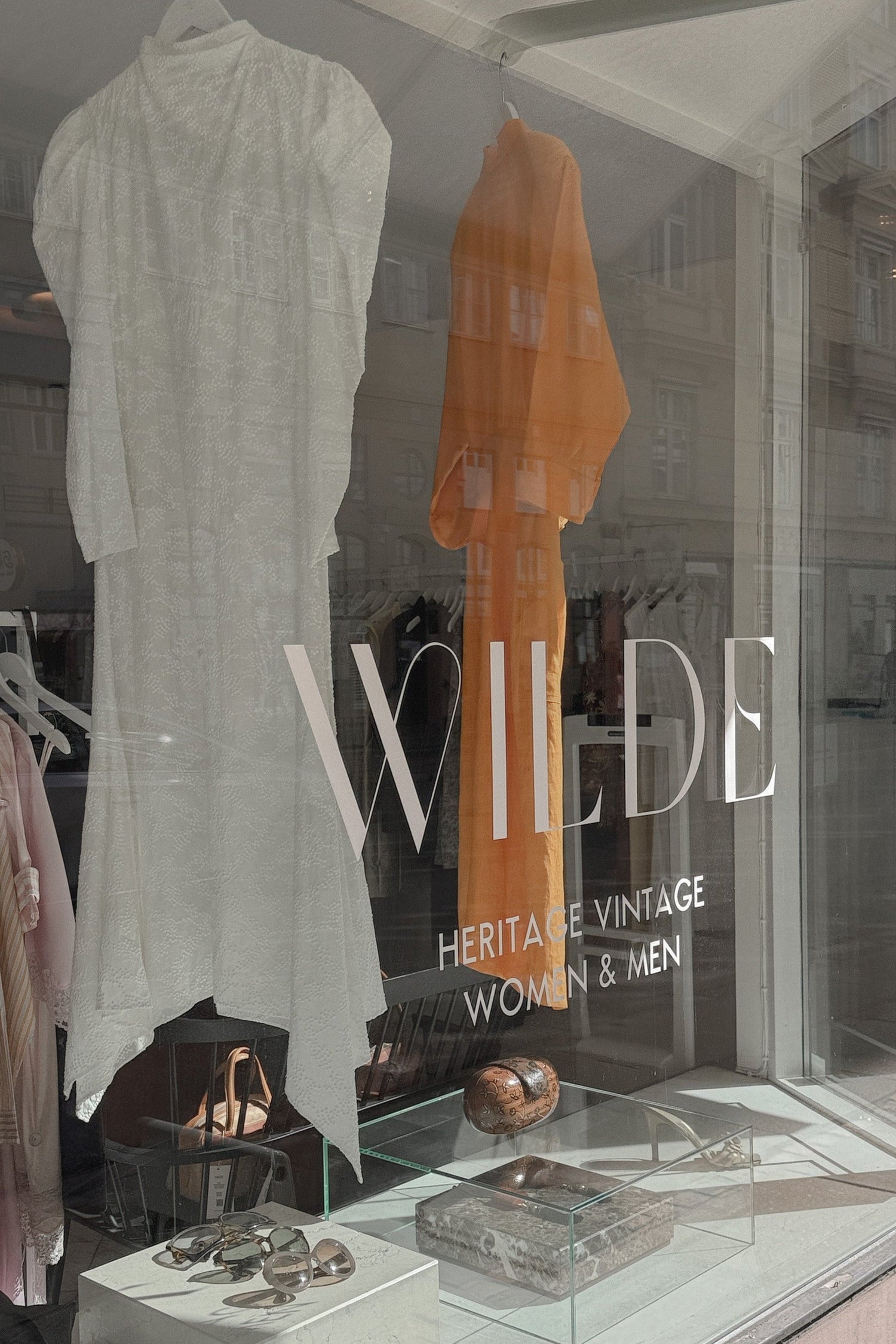 WILDE Copenhagen storefront window display showing fashion items, including a white textured dress, an orange garment, jewelry, and accessories. Large frosted window text reads 'WILDE' with smaller text 'HERITAGE VINTAGE WOMEN & MEN'.